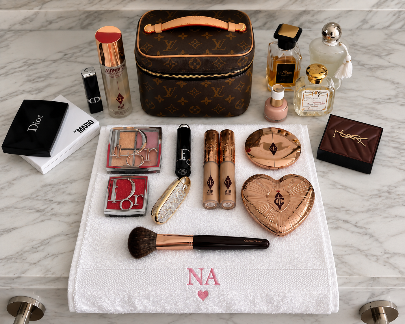 A luxury makeup vanity set arranged on a white marble surface with a white monogrammed towel. The set includes a brown monogram cosmetic bag, Dior and Chanel cosmetics, Charlotte Tilbury lip products, a gold heart-shaped compact, a YSL perfume bottle, and other beauty items.