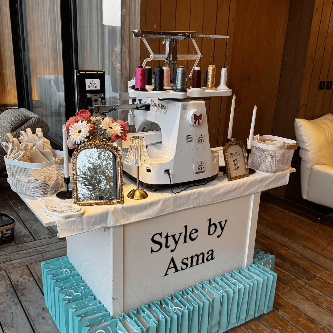 A styled display table in a boutique setting with a white sewing machine, spools of thread, flowers, and many turquoise gift bags arranged around the base. The front of the table reads 'Style by Asma'.